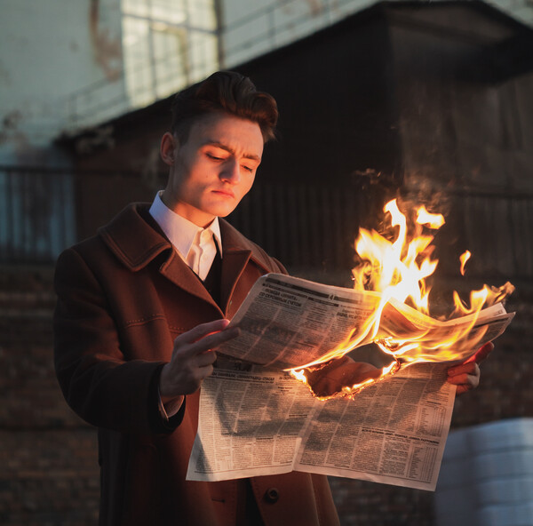 Man standing outside a building and reading a newspaper that's in flames.